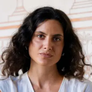 A woman with shoulder-length, curly dark hair looks at the camera with a calm expression and her arms folded. She wears a white sleeveless V-neck top. Background of a white wall with decorative terracotta-coloured line paintings.